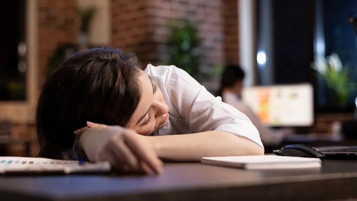 Constant fatigue at work may be more than stress. It could signal a vitamin B12 deficiency in women. Young woman sleeping at her desk in an office, showing signs of fatigue