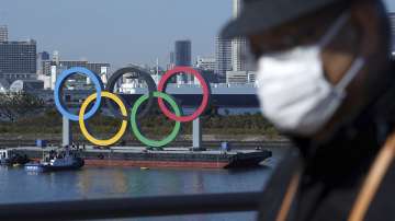 In this Tuesday, Dec. 1, 2020 file photo, a man wearing a protective face mask to help curb the spread of the coronavirus walks with the Olympic rings in the background in the Odaiba section, in Tokyo.
