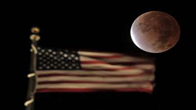 The earth's shadow covers the full moon during a partial lunar eclipse as it sets beyond the U.S. flag on top of a building, Friday, Nov. 19, 2021, in downtown Kansas City, Missouri.